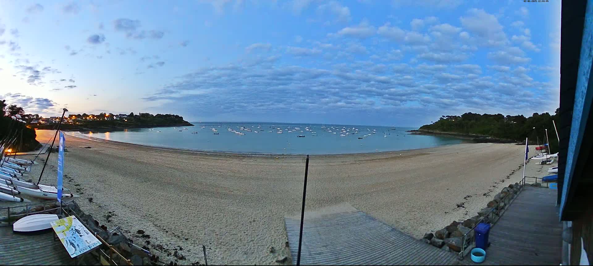 A sandy beach curves around a calm bay filled with numerous small boats under a partly cloudy, twilight sky.
