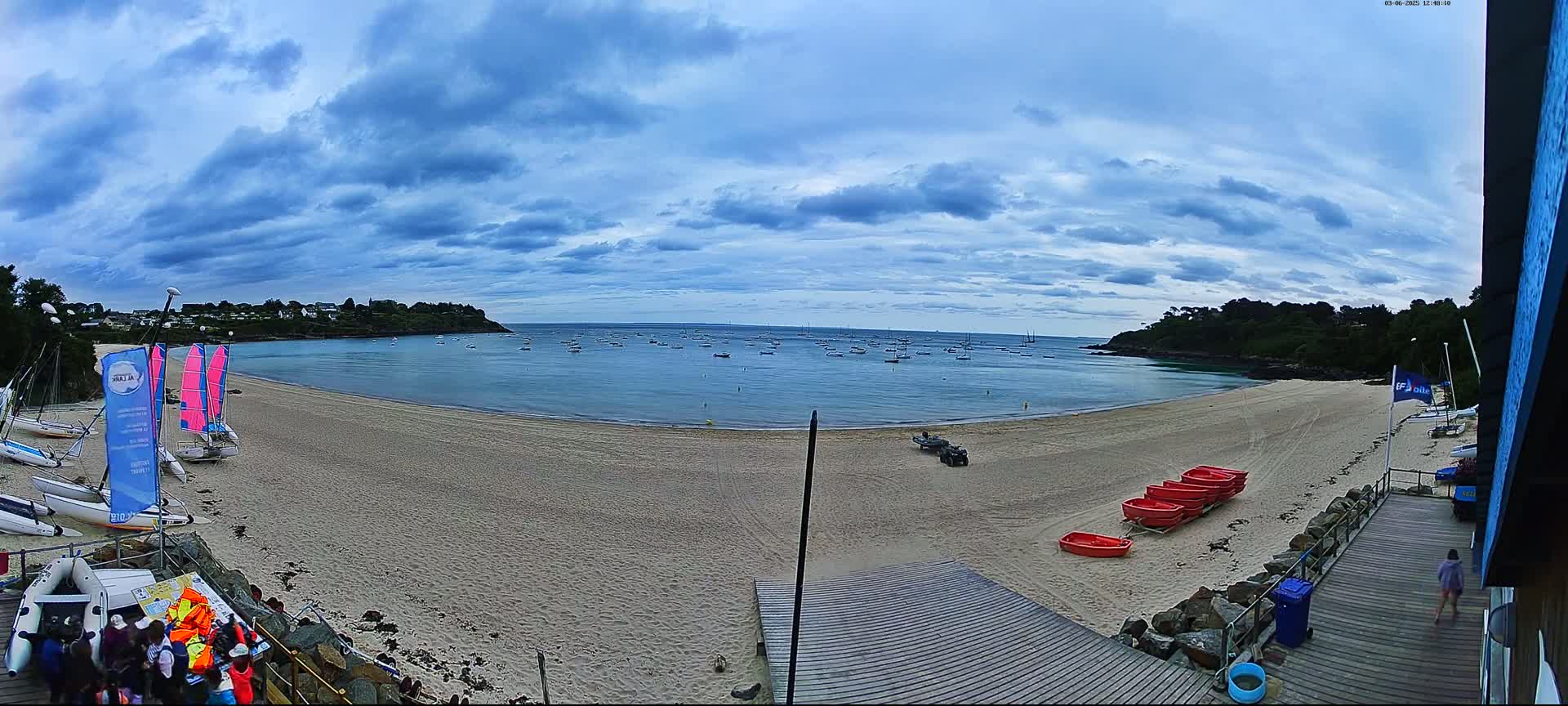 A sandy beach with sailboats in the water and several red rescue boats on the shore is under a partly cloudy sky.