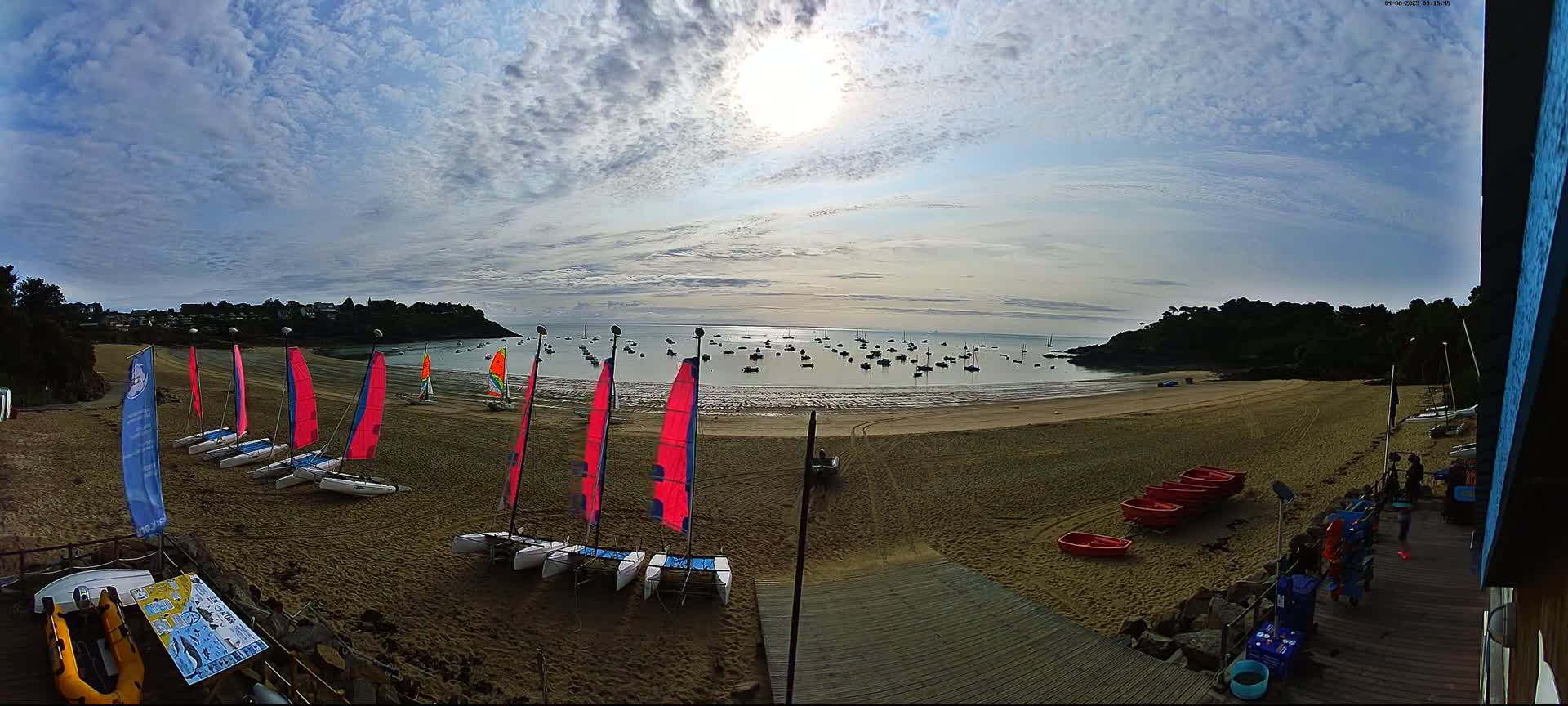 A sandy beach with numerous sailboats on the water and several catamarans on the shore under a partly sunny sky.