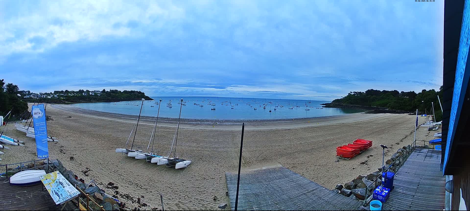 A sandy beach with several sailboats on the shore and many more boats in the calm water of a bay under a partly cloudy sky.