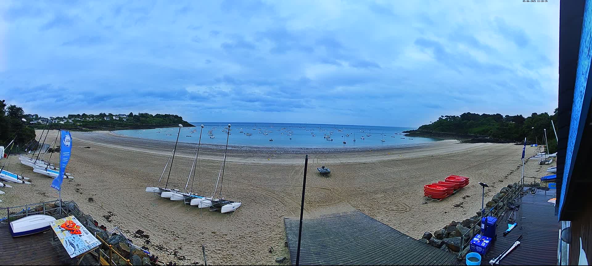 A sandy beach with several sailboats on the shore and many more in the calm water under a cloudy sky.