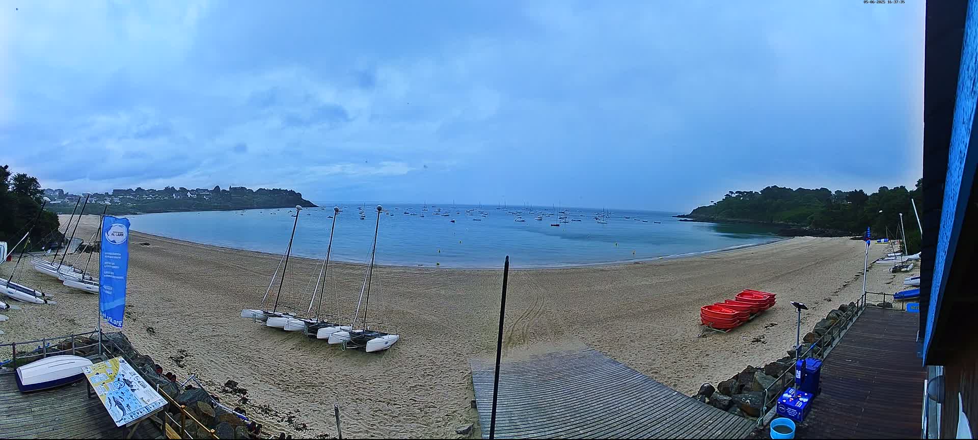 A sandy beach with several sailboats on the shore and more boats in the calm, cloudy water, is nestled between green hills under a mostly cloudy sky.