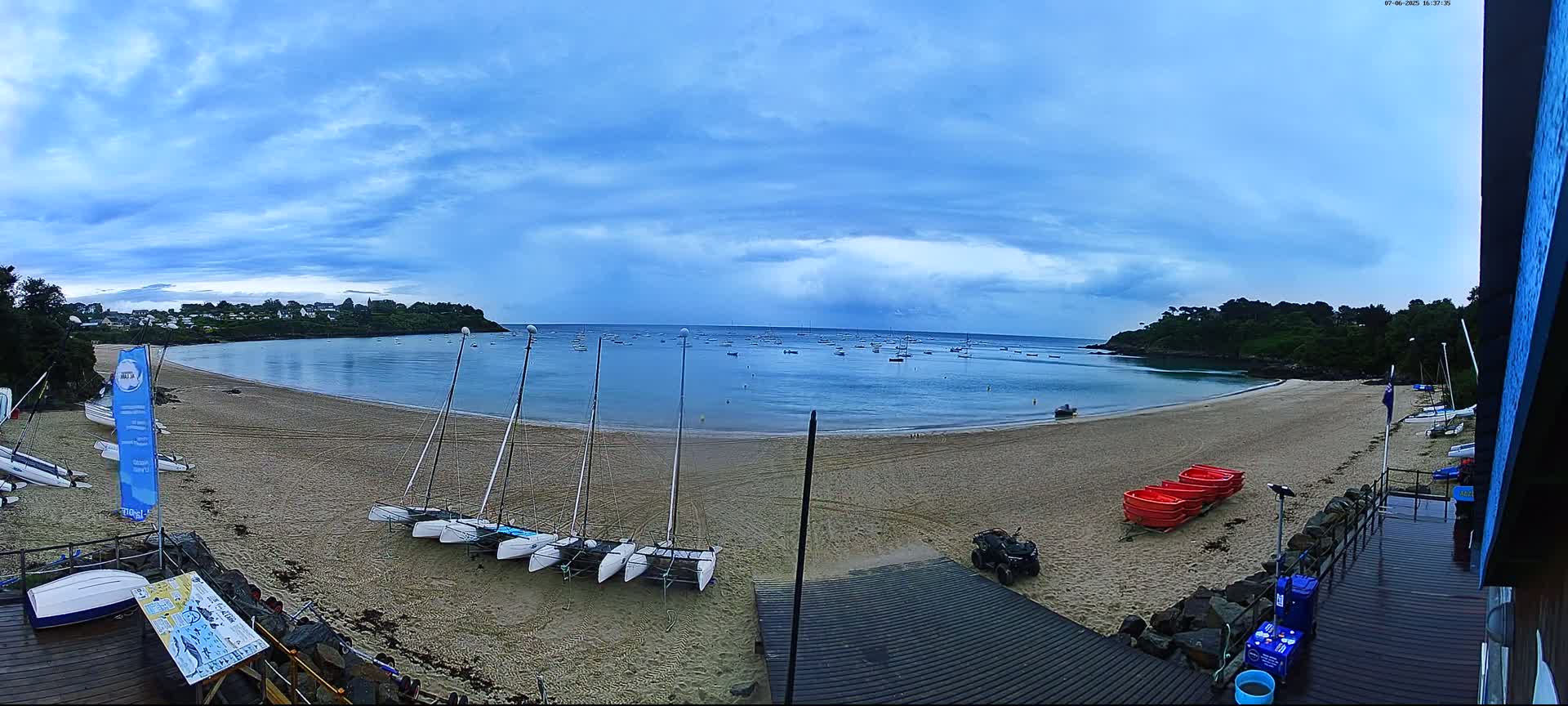 A sandy beach with several sailboats on the shore and numerous boats in the calm bay under a partly cloudy sky.