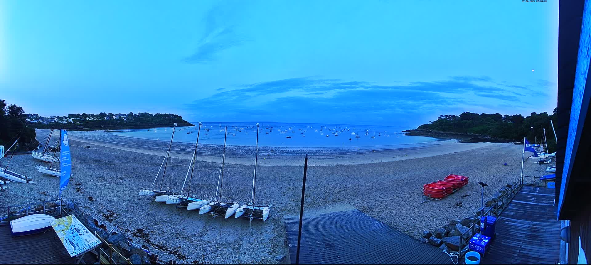 Under a twilight sky, several sailboats are beached on a sandy bay with calm water and numerous boats visible on the horizon.