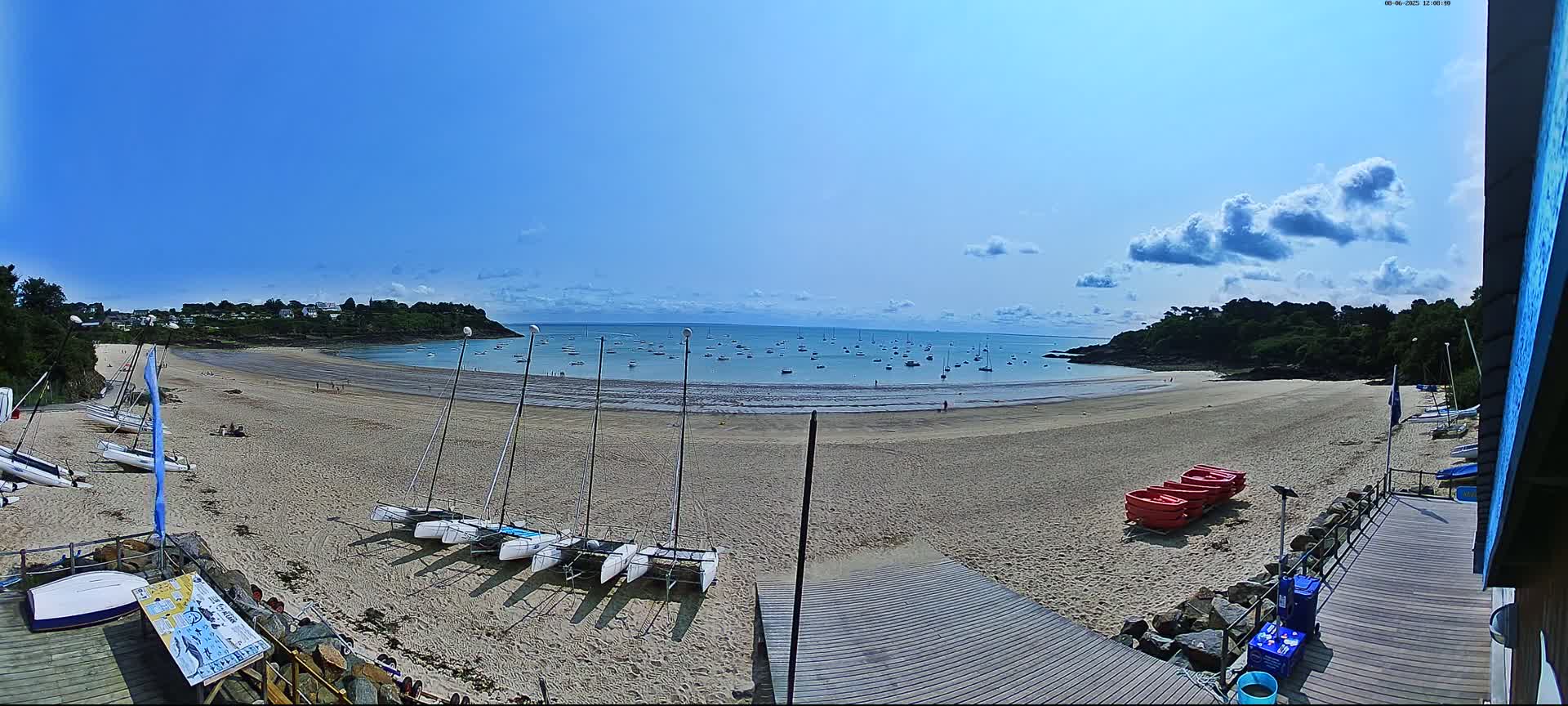 A sandy beach with several sailboats on the shore and many more in the calm, blue water under a mostly sunny sky with some scattered clouds.