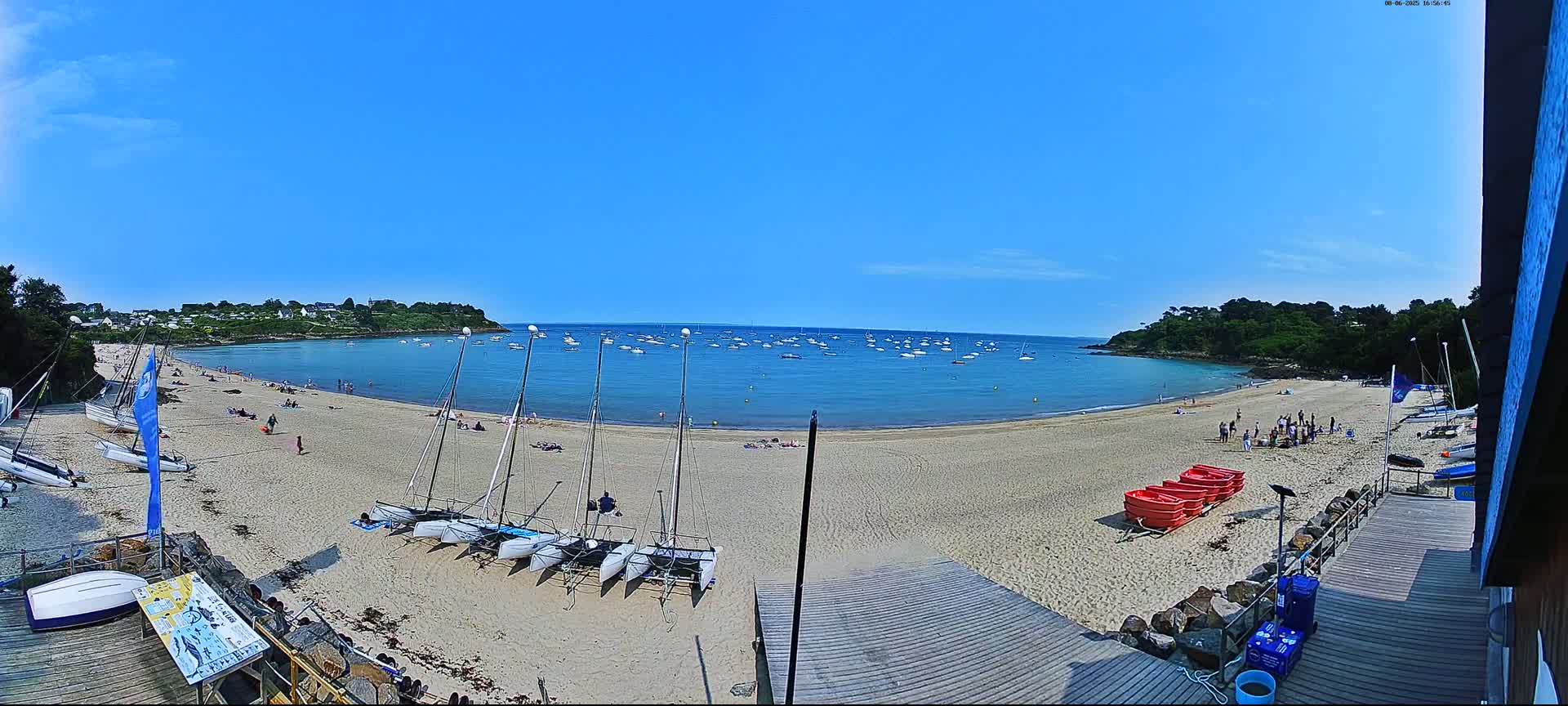 A sandy beach with numerous sailboats on the shore and in the calm, blue water is surrounded by green hills under a clear, sunny sky.