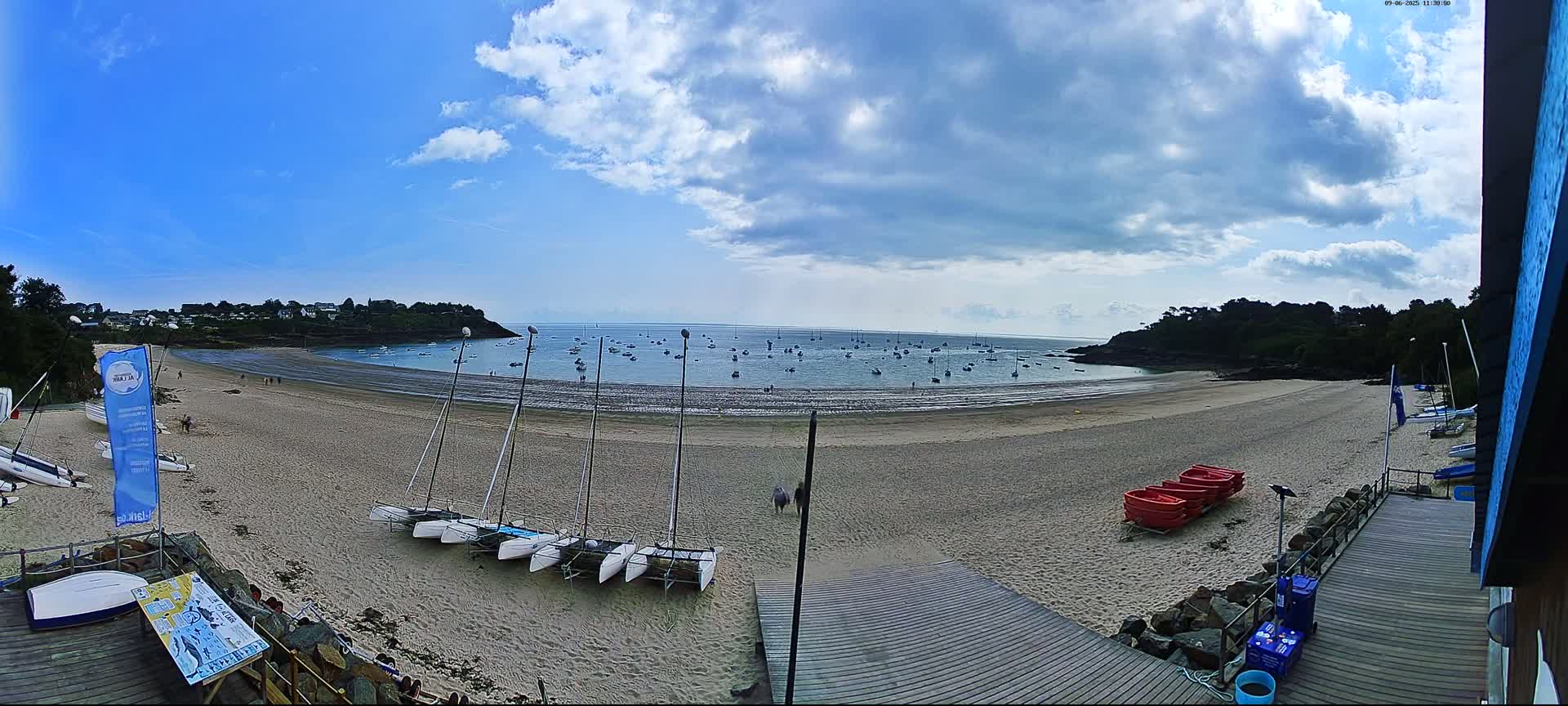 A sandy beach with several sailboats on the shore and many more in the calm water under a partly cloudy blue sky.