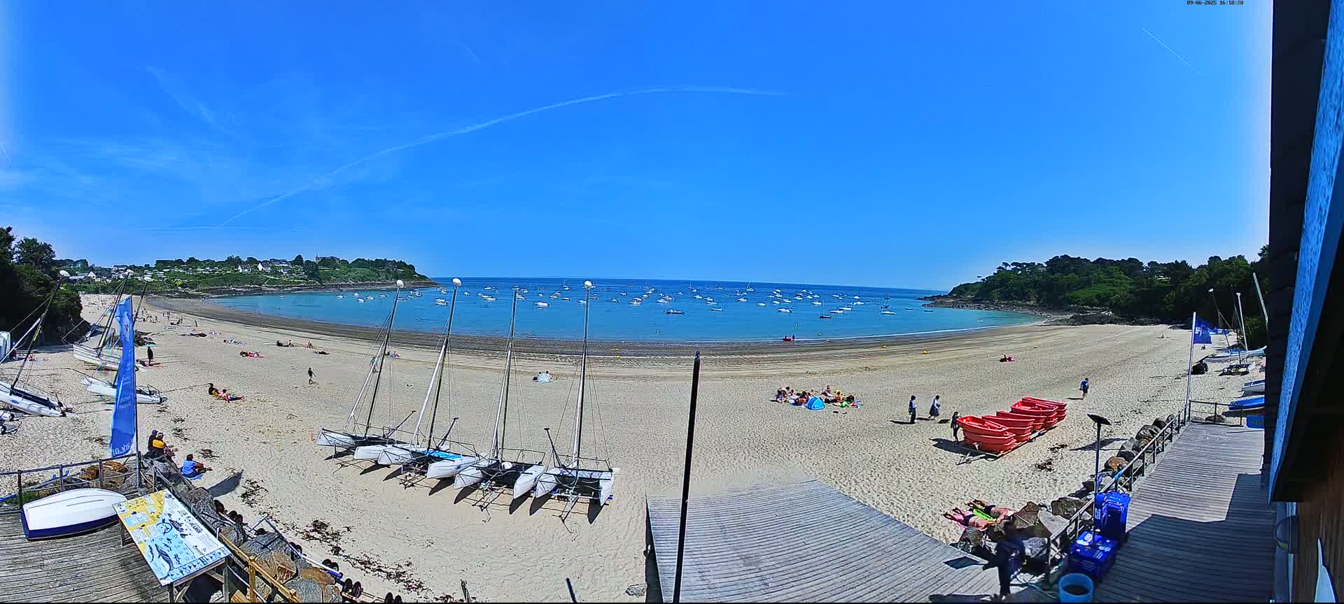 A sandy beach with numerous people sunbathing and several sailboats on the shore and in the calm, clear blue water under a bright, sunny sky.