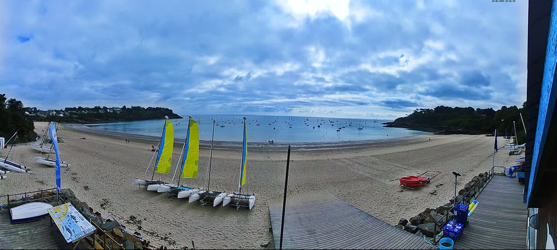 A sandy beach with several sailboats on the shore and many more in the calm bay under a partly cloudy sky.