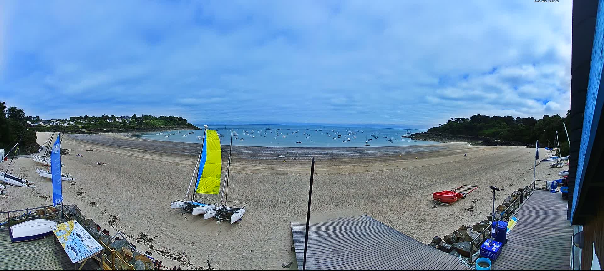 A sandy beach curves around a calm bay dotted with sailboats under a partly cloudy sky, with several catamarans beached nearby.