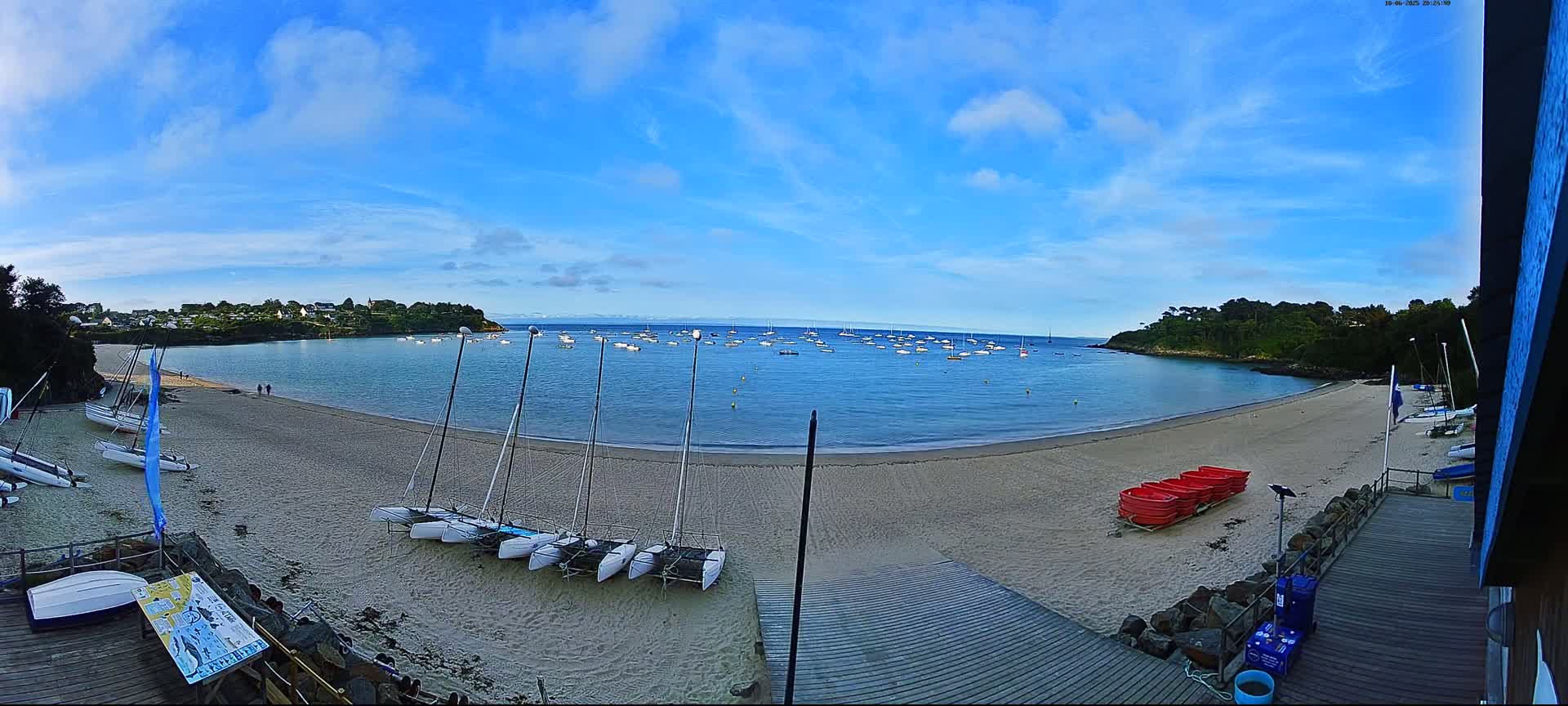 A sandy beach with several sailboats on the shore and many more in the calm, blue water under a mostly clear, blue sky.