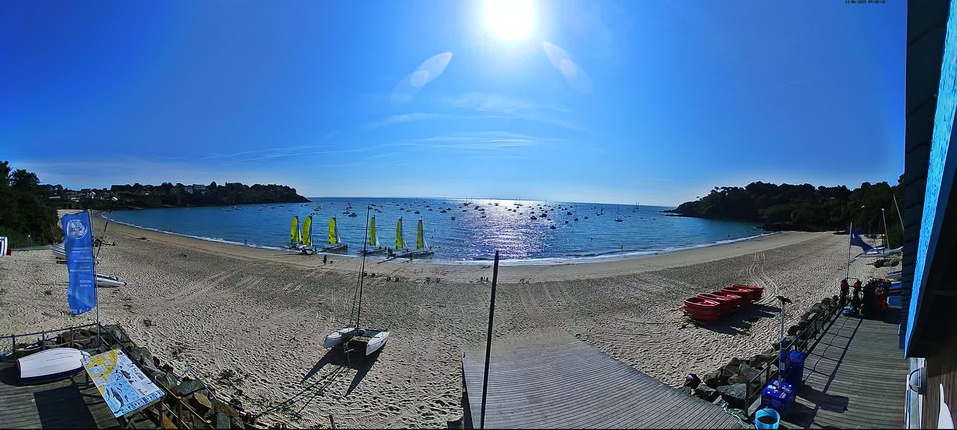 A sandy beach with several sailboats on the water and more beached on the sand, under a bright sunny sky.