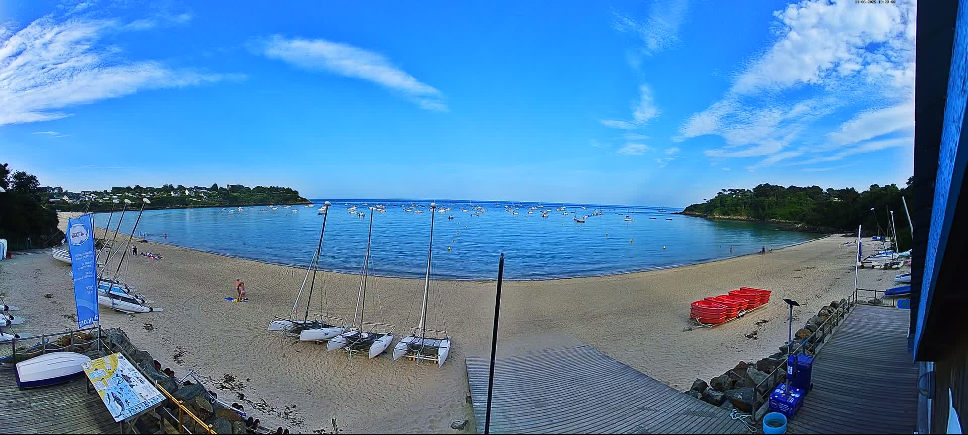 A sandy beach with several sailboats and small boats on the shore and in the calm, blue water under a mostly sunny sky with a few scattered clouds.