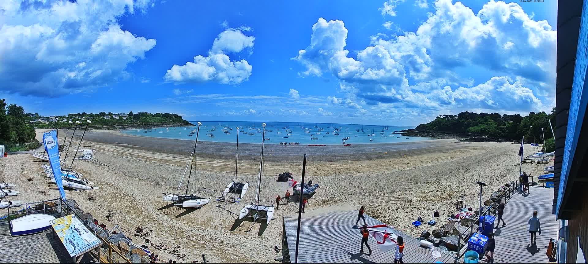 A sandy beach with numerous sailboats on the shore and in the calm water under a partly cloudy sky.