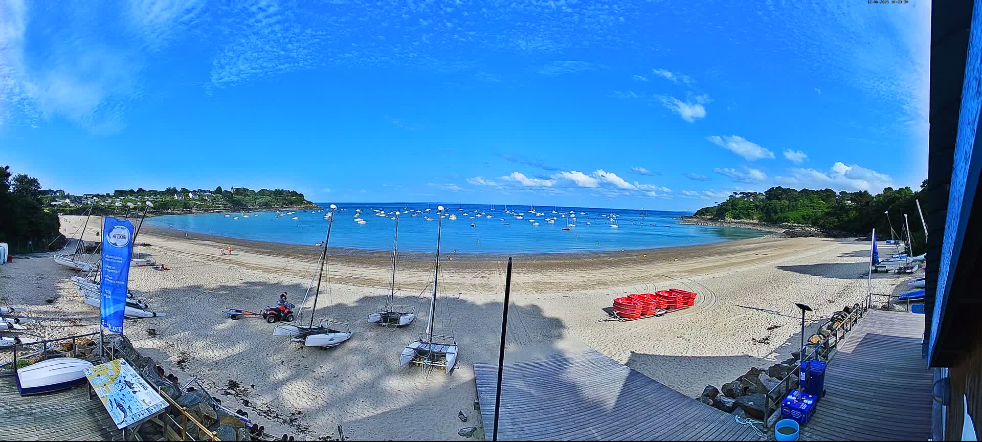 A sandy beach curves around a calm bay dotted with sailboats under a mostly sunny sky.