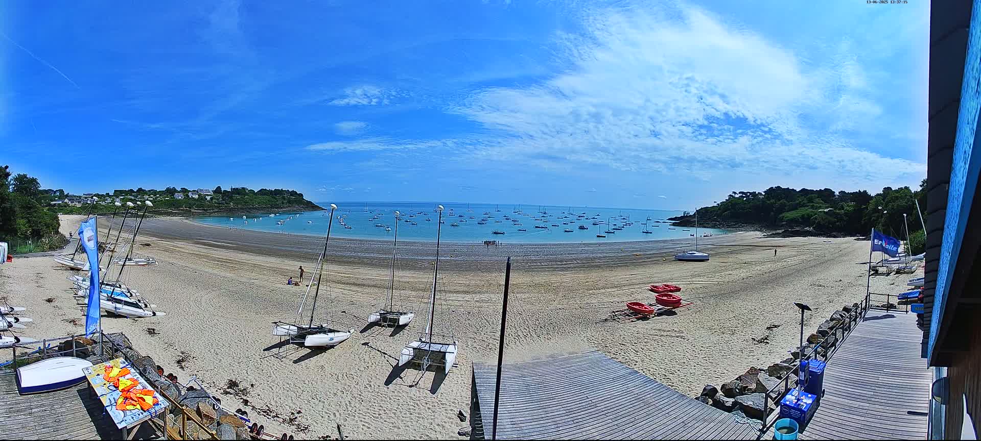 A sandy beach with numerous sailboats on the shore and in the calm water under a mostly sunny sky.