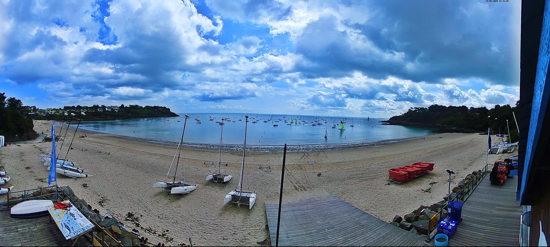 A sandy beach with numerous sailboats on the water and several catamarans beached, under a partly cloudy sky.