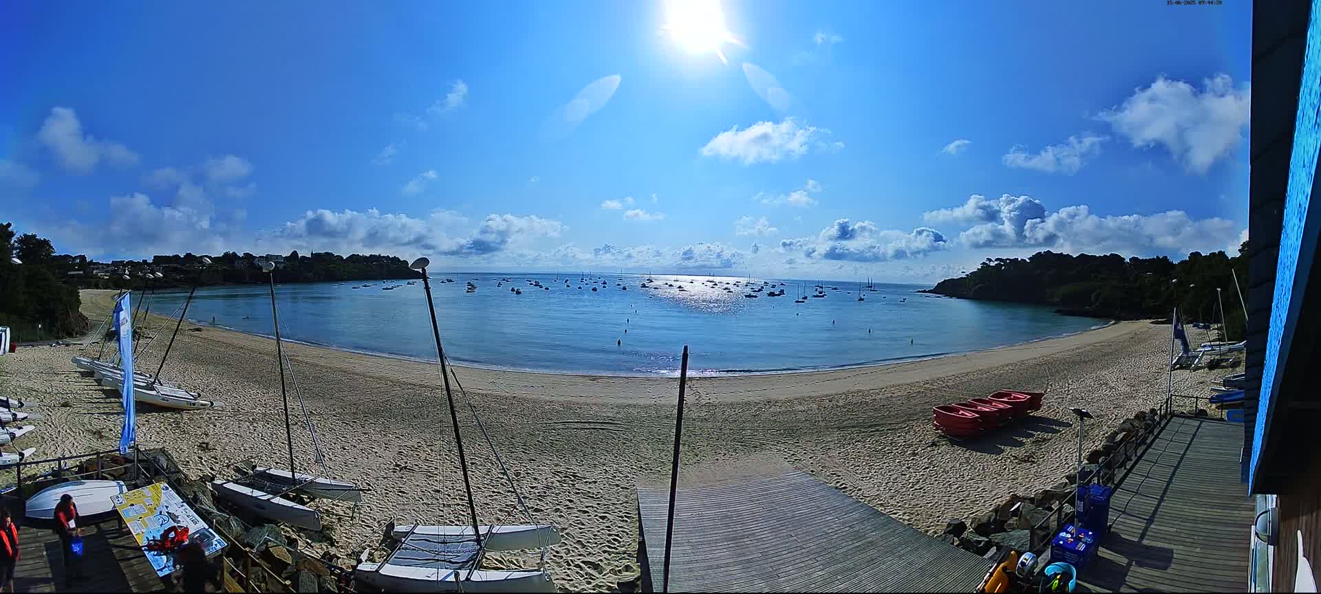 A sandy beach curves around a calm, sunny bay dotted with sailboats, under a mostly clear blue sky with scattered clouds.