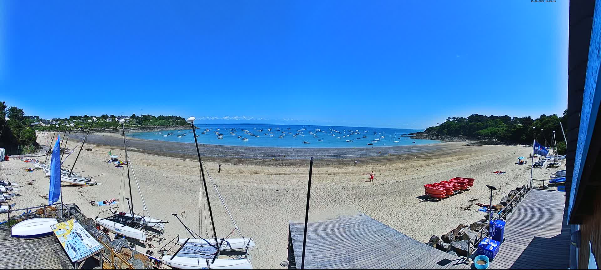 A sandy beach with numerous sailboats on the shore and in the calm, blue water under a clear, sunny sky.