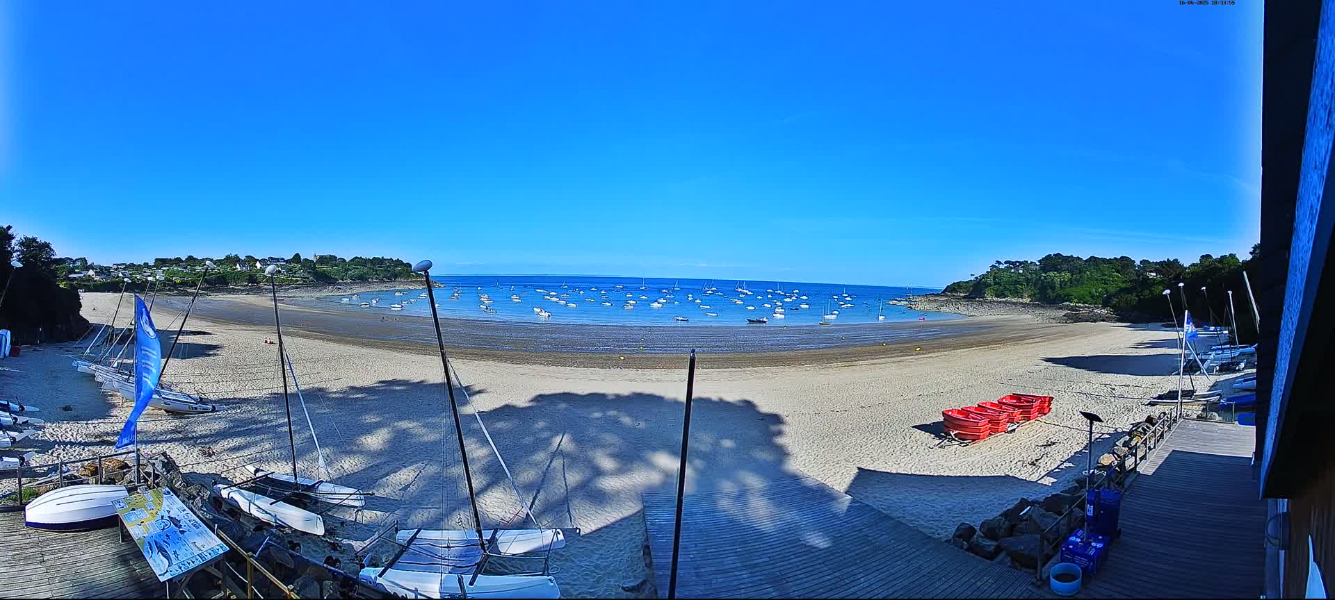 A sandy beach with numerous sailboats in the calm water under a clear blue sky, and several sailboats and red rowboats on the shore.