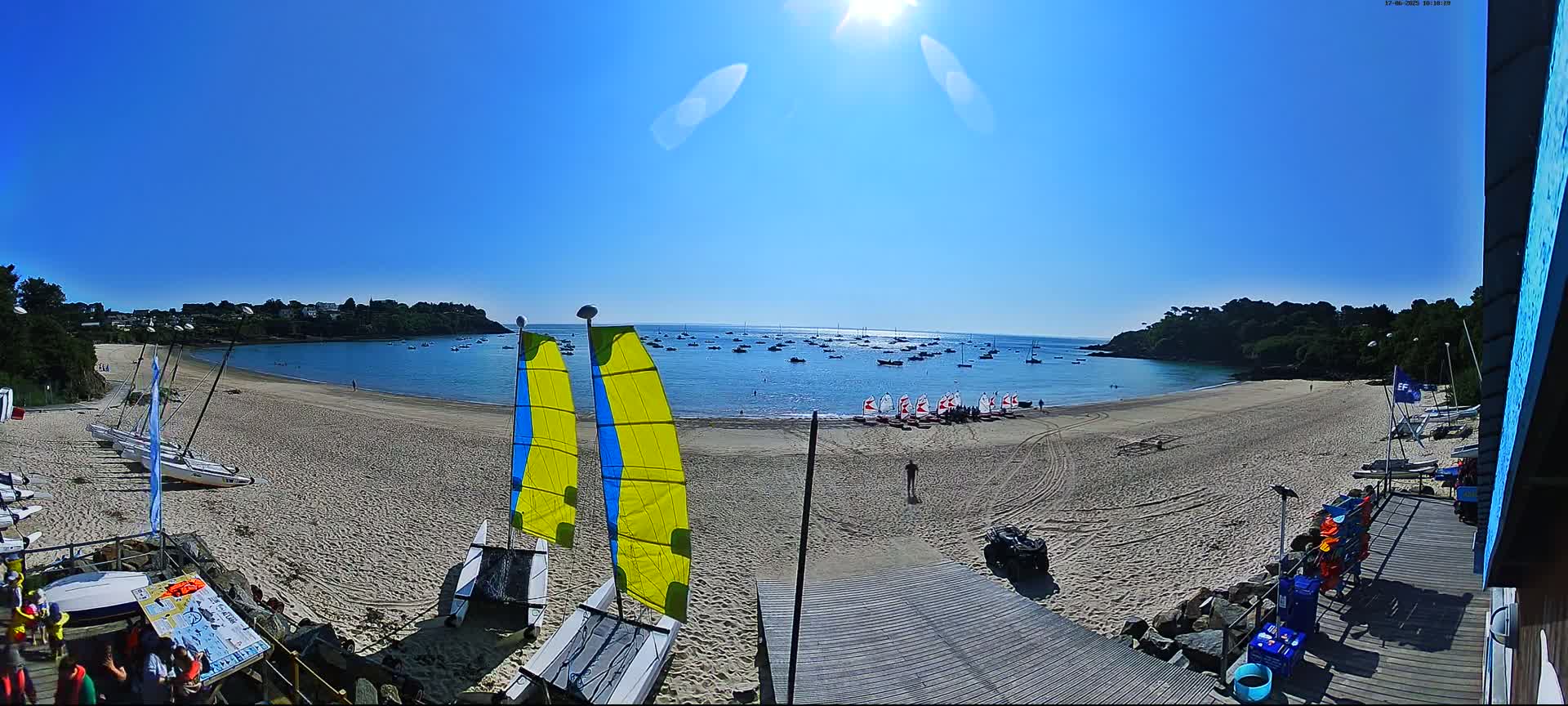 A sandy beach with numerous sailboats on the calm water and several small sailboats on the shore under a clear, sunny sky.