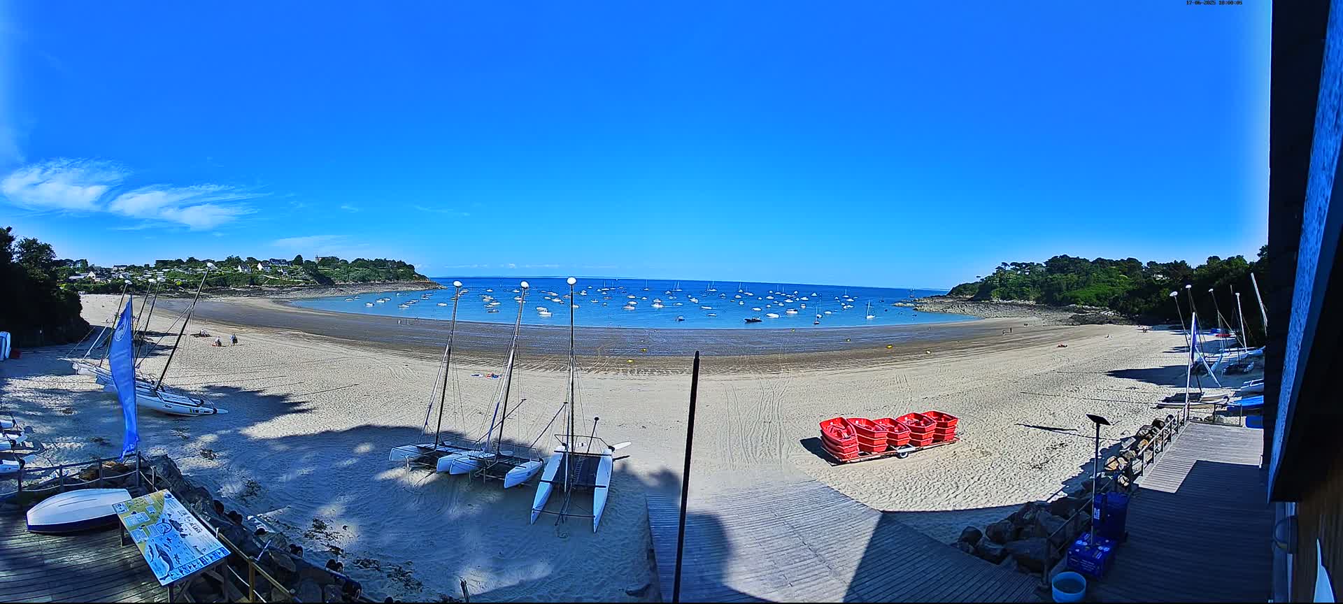 A sandy beach curves around a calm bay under a bright blue sky, with numerous sailboats in the water and several catamarans beached on the sand.