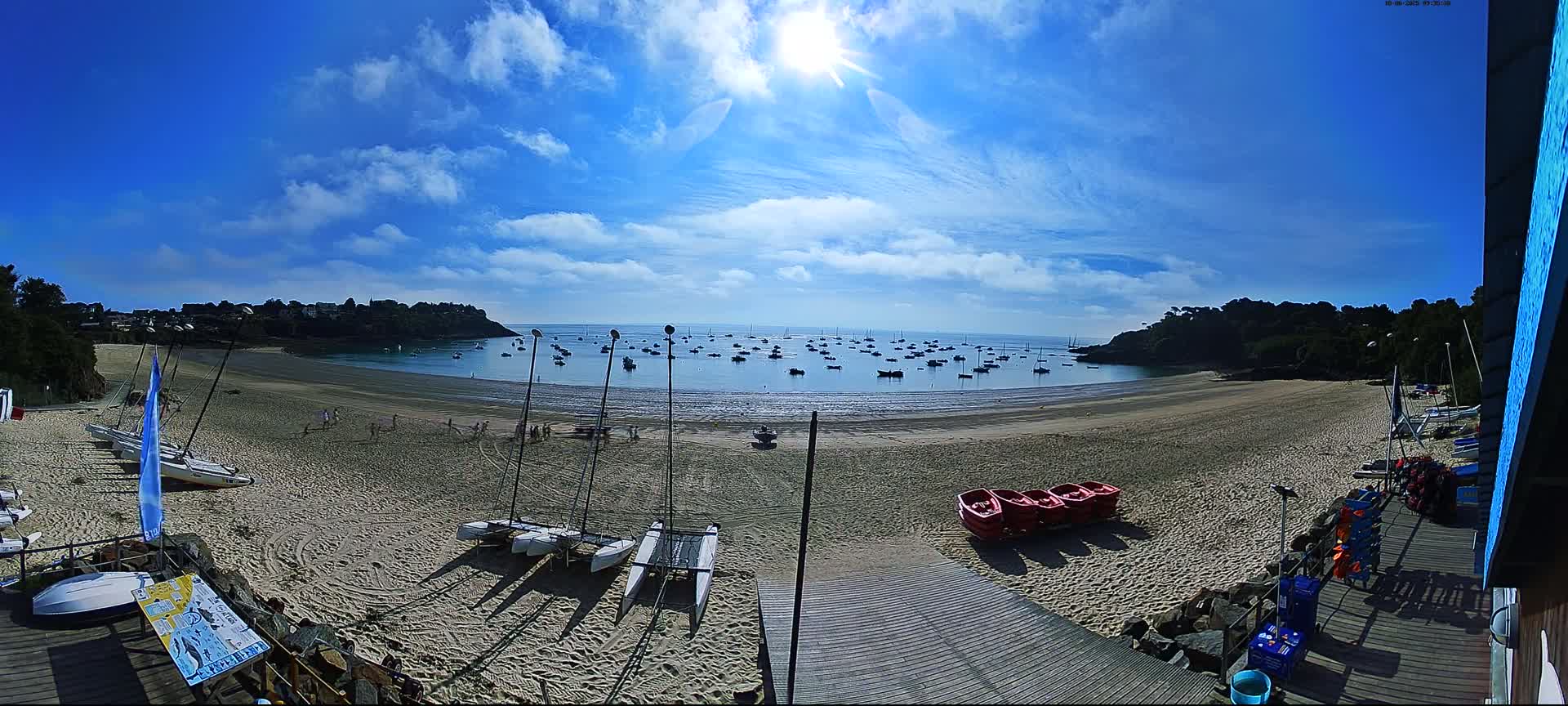 A sandy beach with sailboats on the shore and numerous boats in the calm, sunny water, is surrounded by green hills under a bright, mostly clear sky.