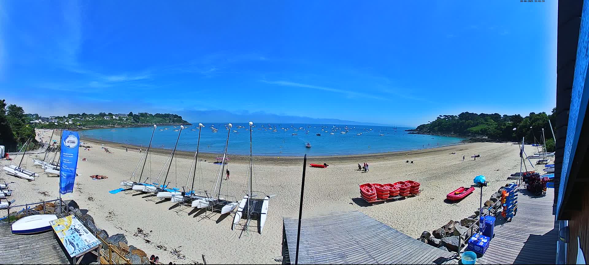 A sandy beach with numerous sailboats on the shore and a calm bay filled with boats under a clear blue sky.