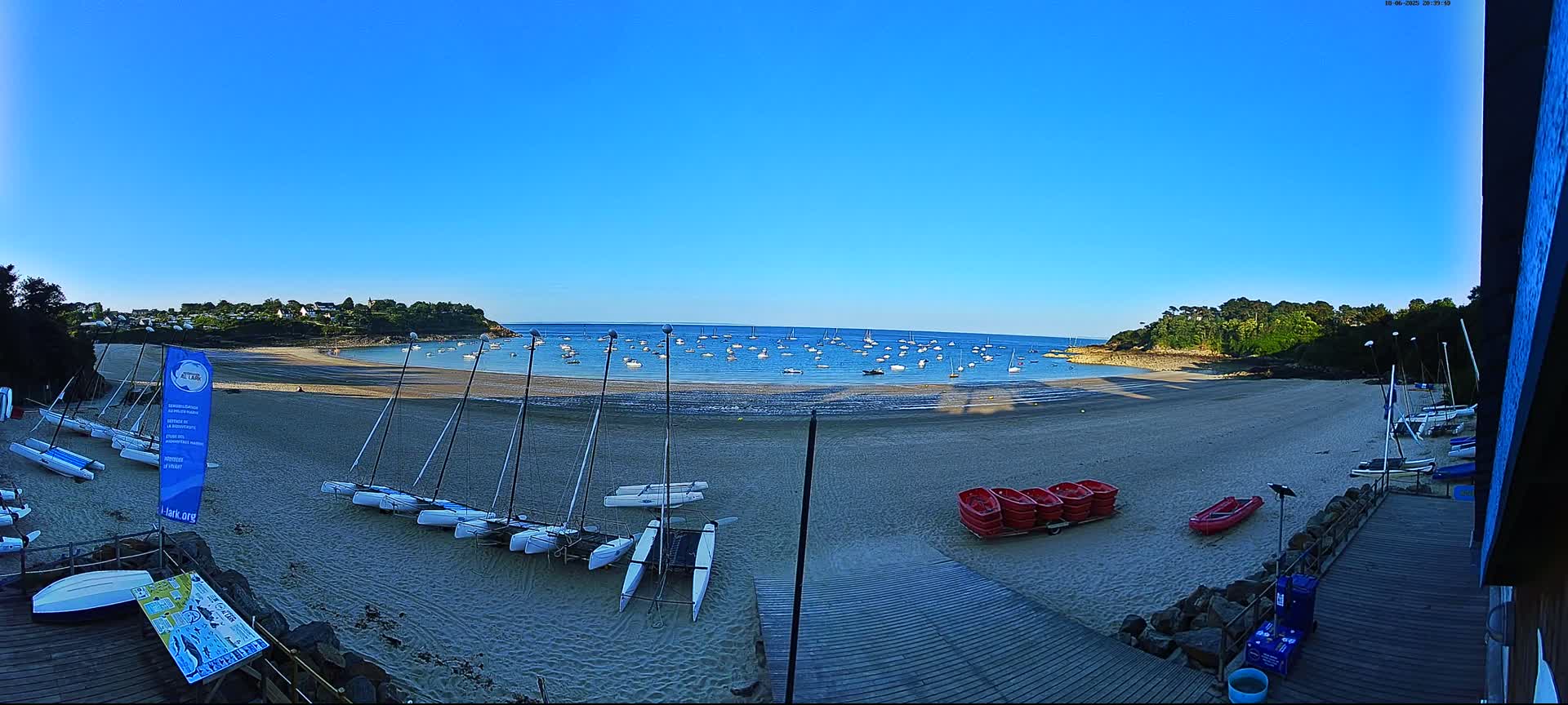 A sandy beach with numerous sailboats on the water and several catamarans on the shore under a clear blue sky.