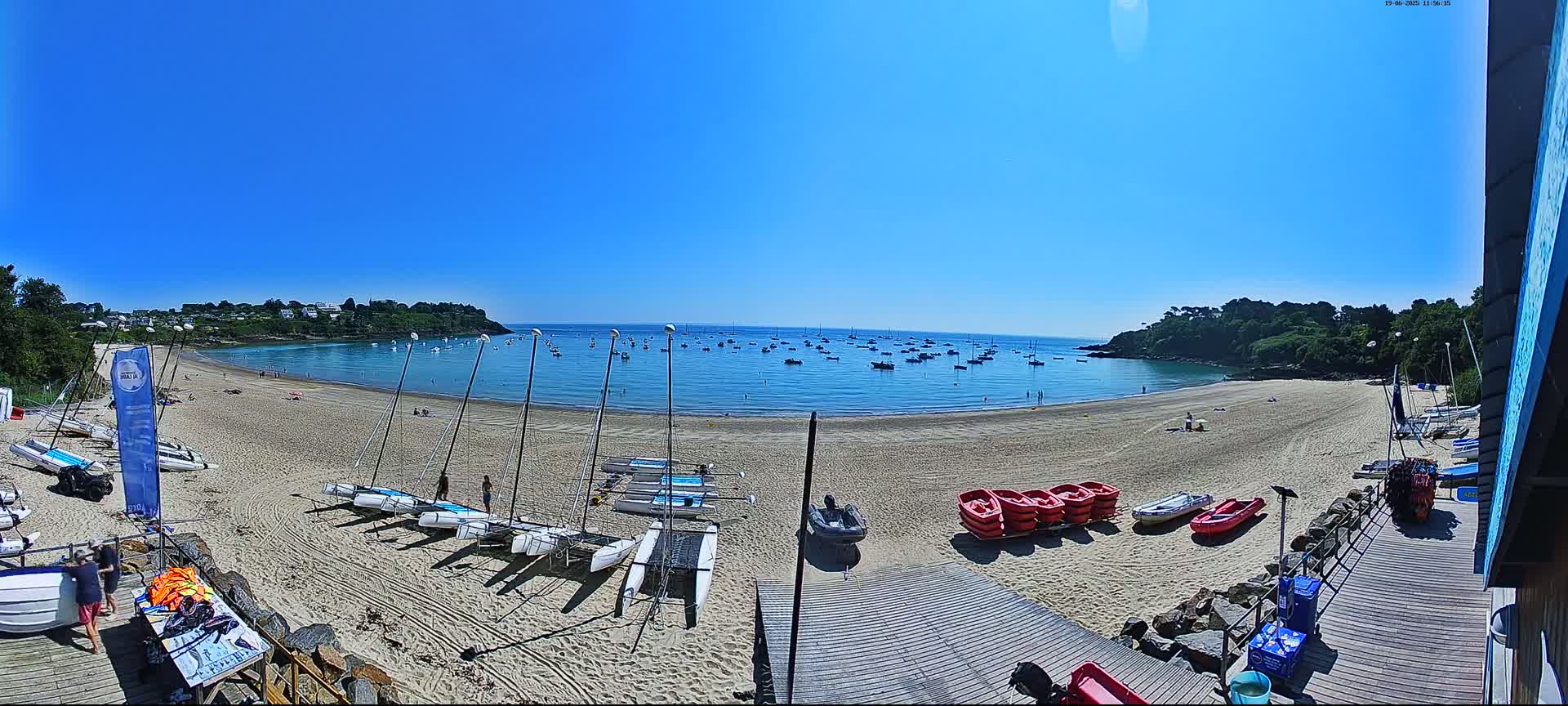 A sandy beach with numerous sailboats and small motorboats on the shore, and a calm bay with more boats under a clear blue sky.