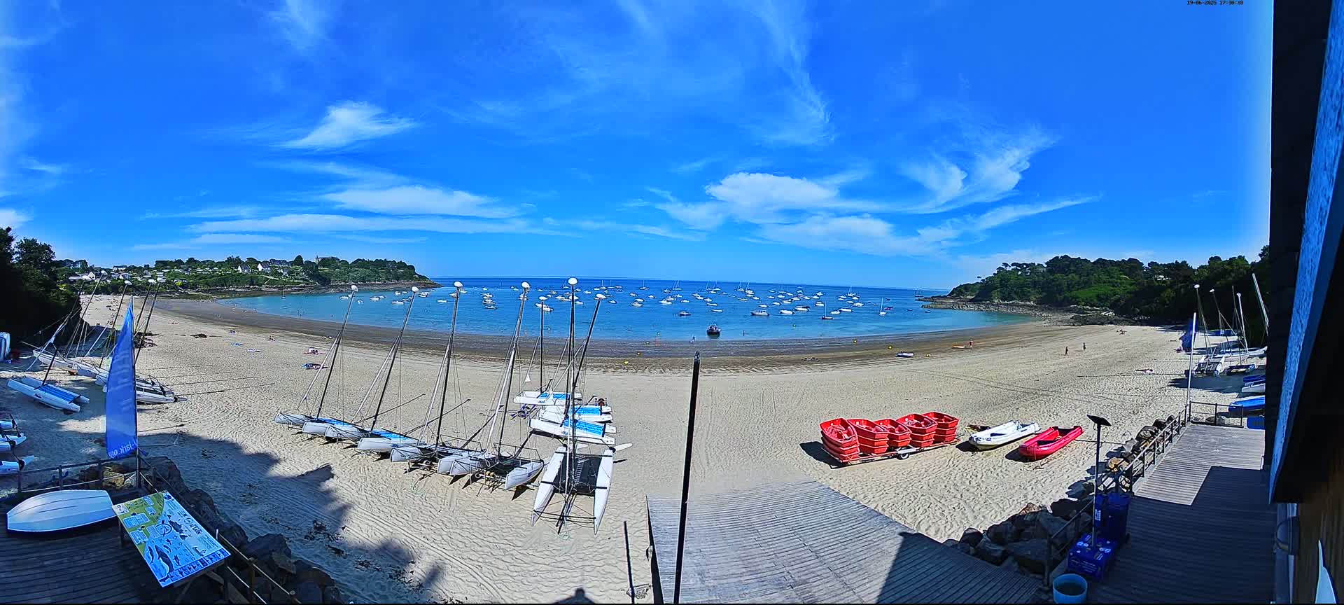 A sandy beach with numerous sailboats on the shore and in the calm, blue water under a mostly sunny sky with scattered clouds.