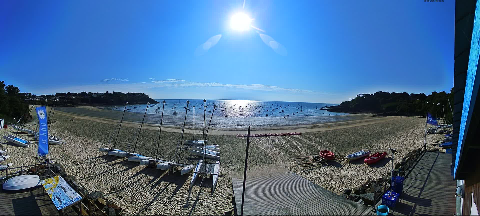 A sandy beach with numerous sailboats on the shore and in the calm, sunlit water is bordered by green hills under a clear blue sky.