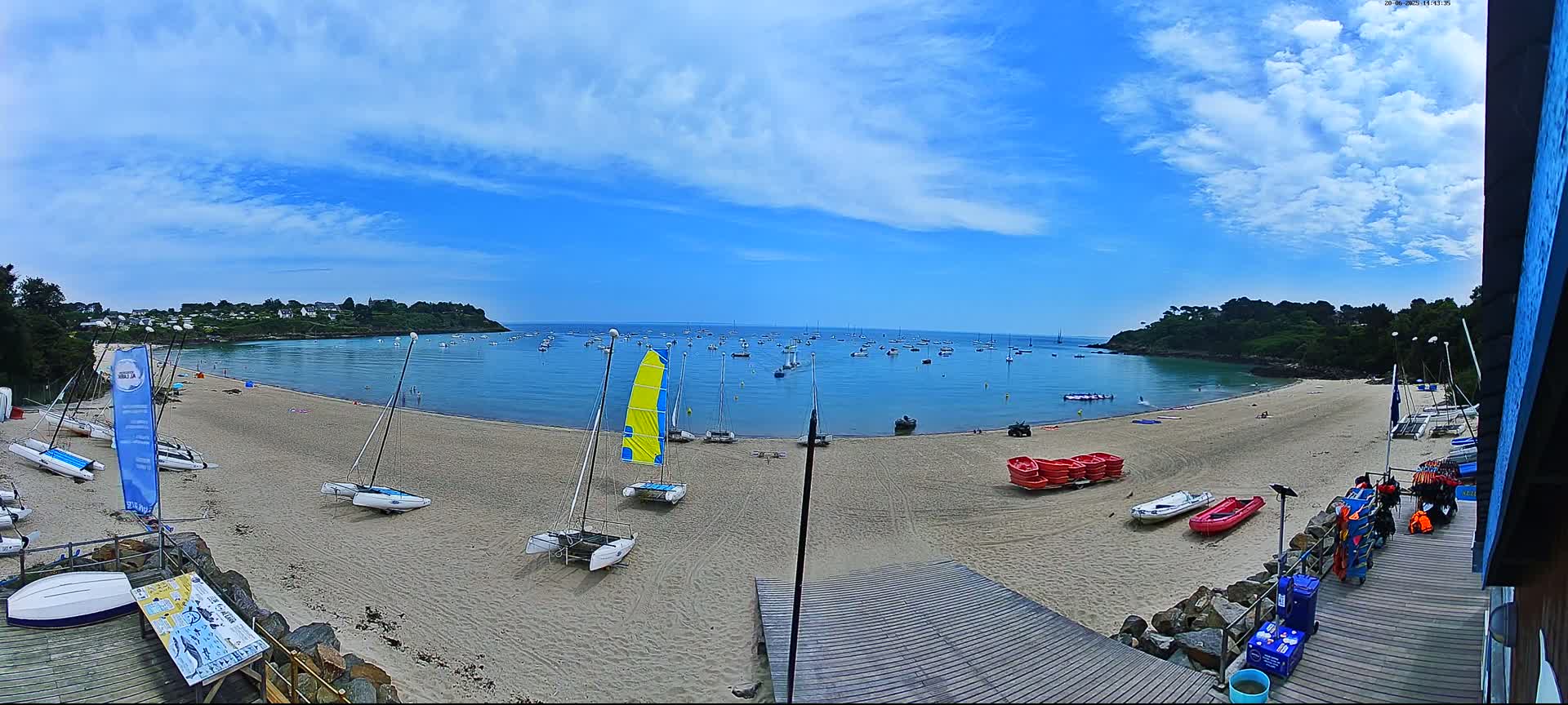 A sandy beach with numerous sailboats on the shore and in the calm bay under a partly cloudy blue sky.