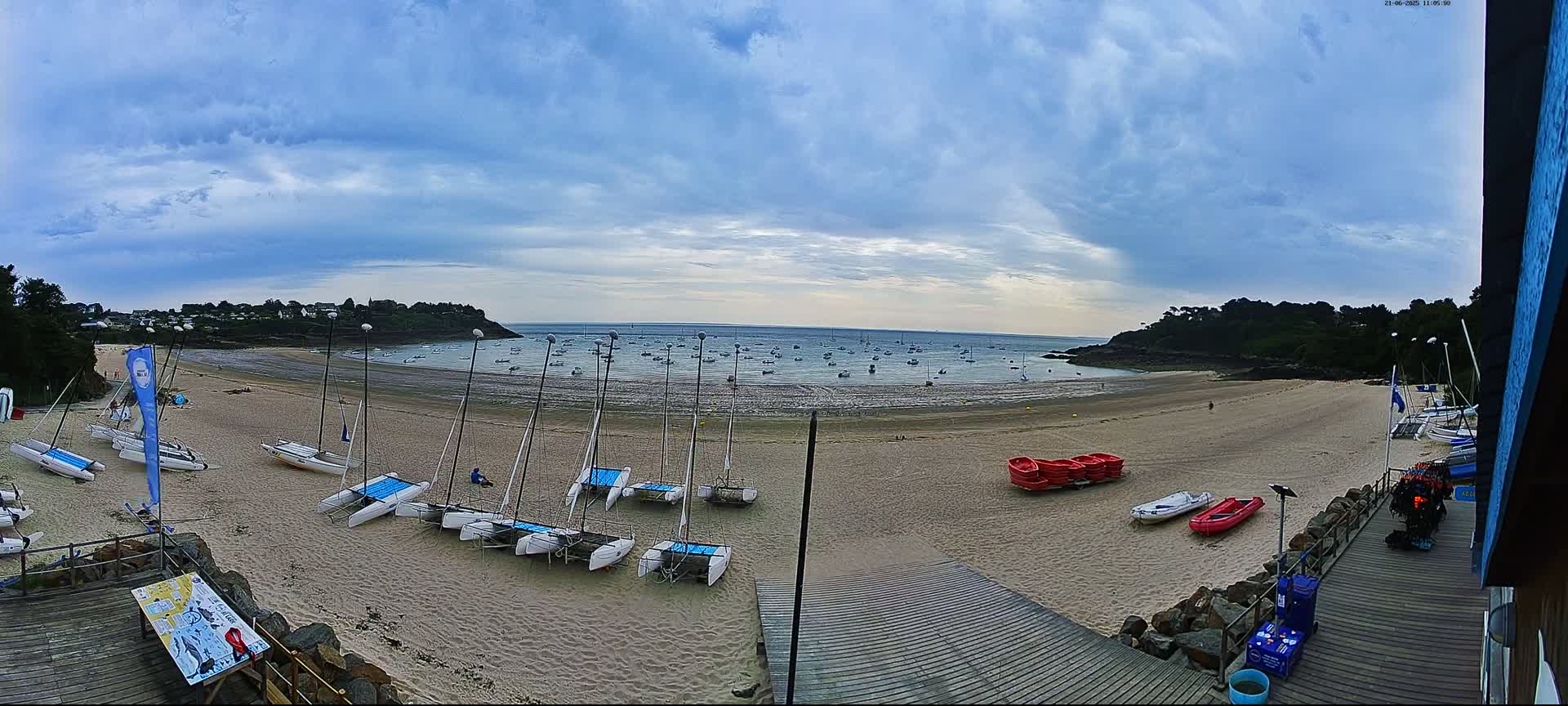 A sandy beach with numerous sailboats and small watercraft on the shore, under a partly cloudy sky, overlooks a calm bay dotted with more boats.