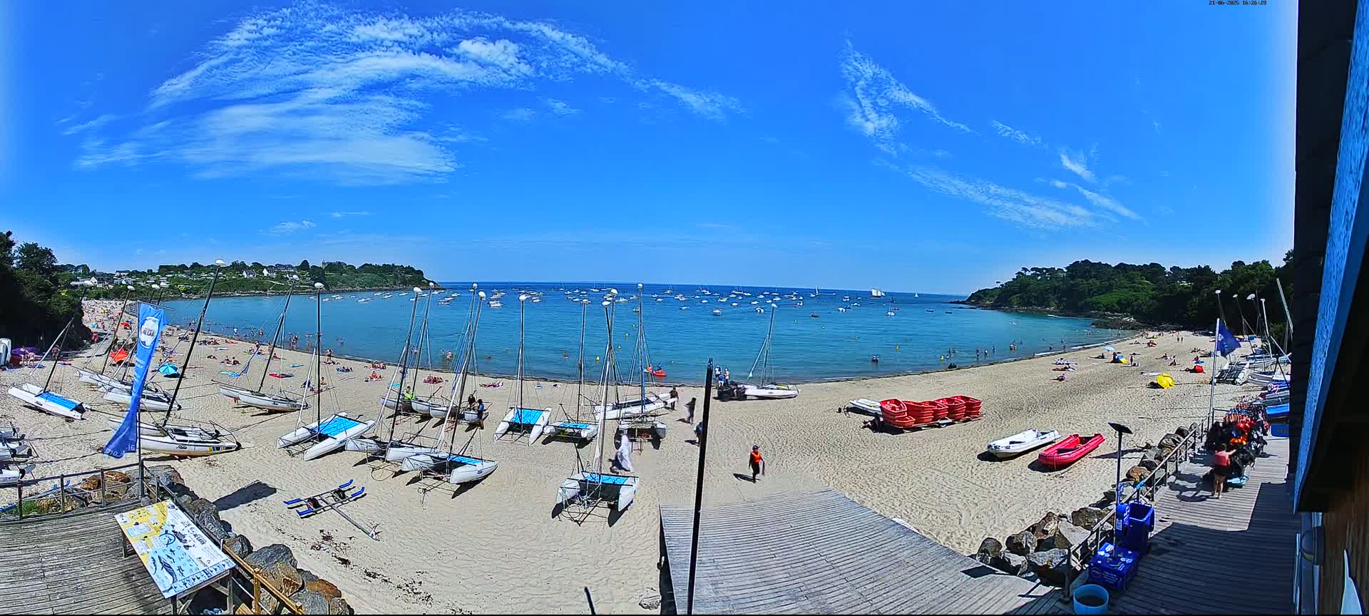 A sandy beach with numerous sailboats and people, under a bright sunny sky with scattered clouds, overlooks a calm bay filled with more boats.