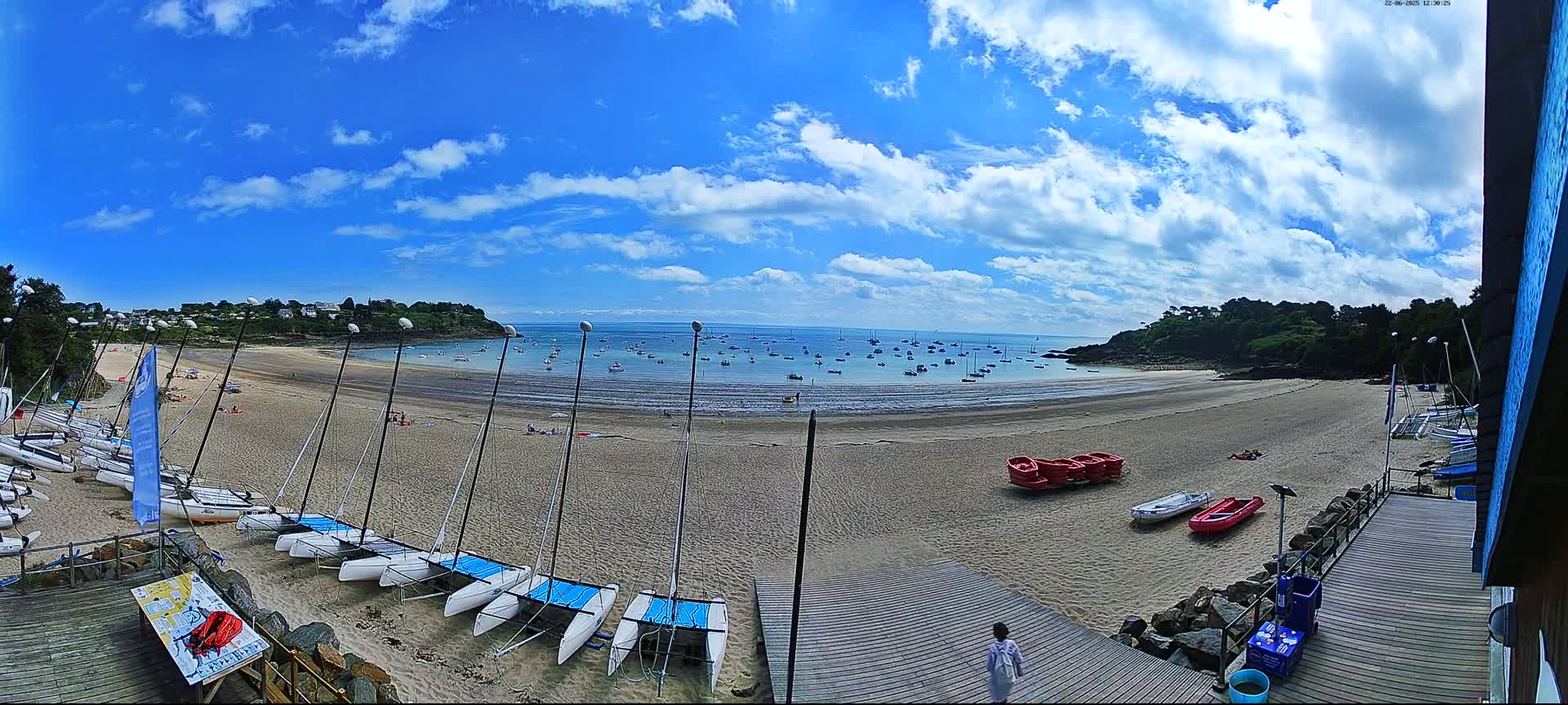 A sandy beach with numerous sailboats and small boats on the shore, under a partly cloudy blue sky, overlooks a calm bay dotted with more boats.