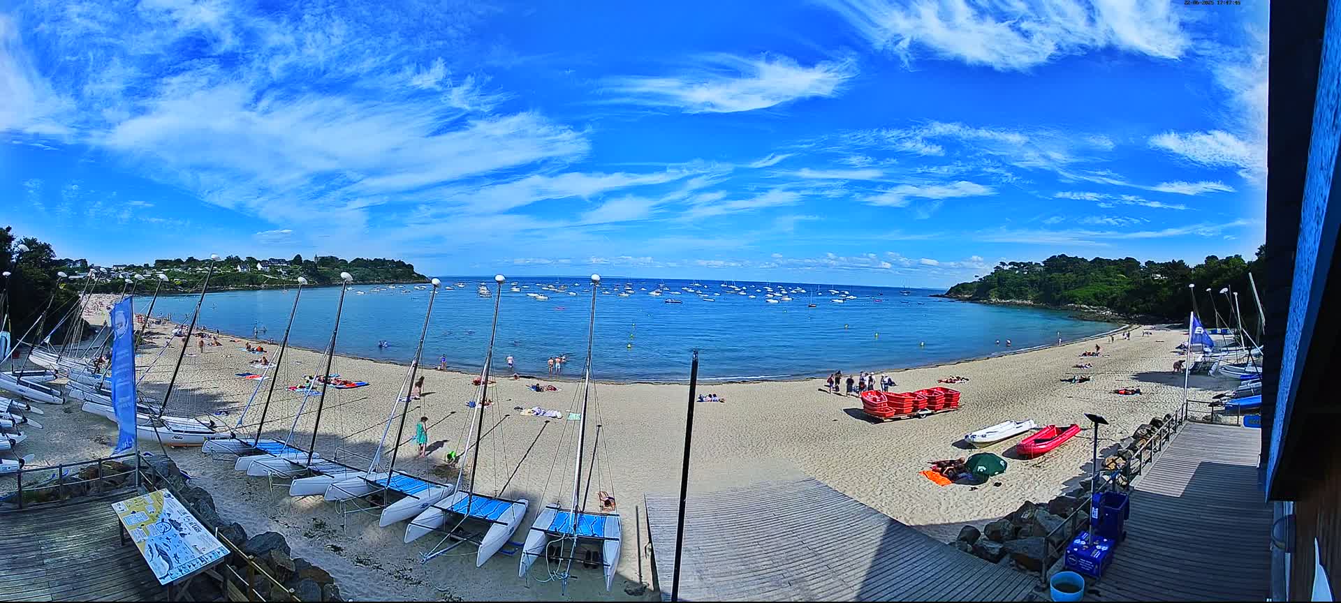 A sandy beach with numerous sailboats and people enjoying a sunny day with a bright blue sky filled with wispy clouds.
