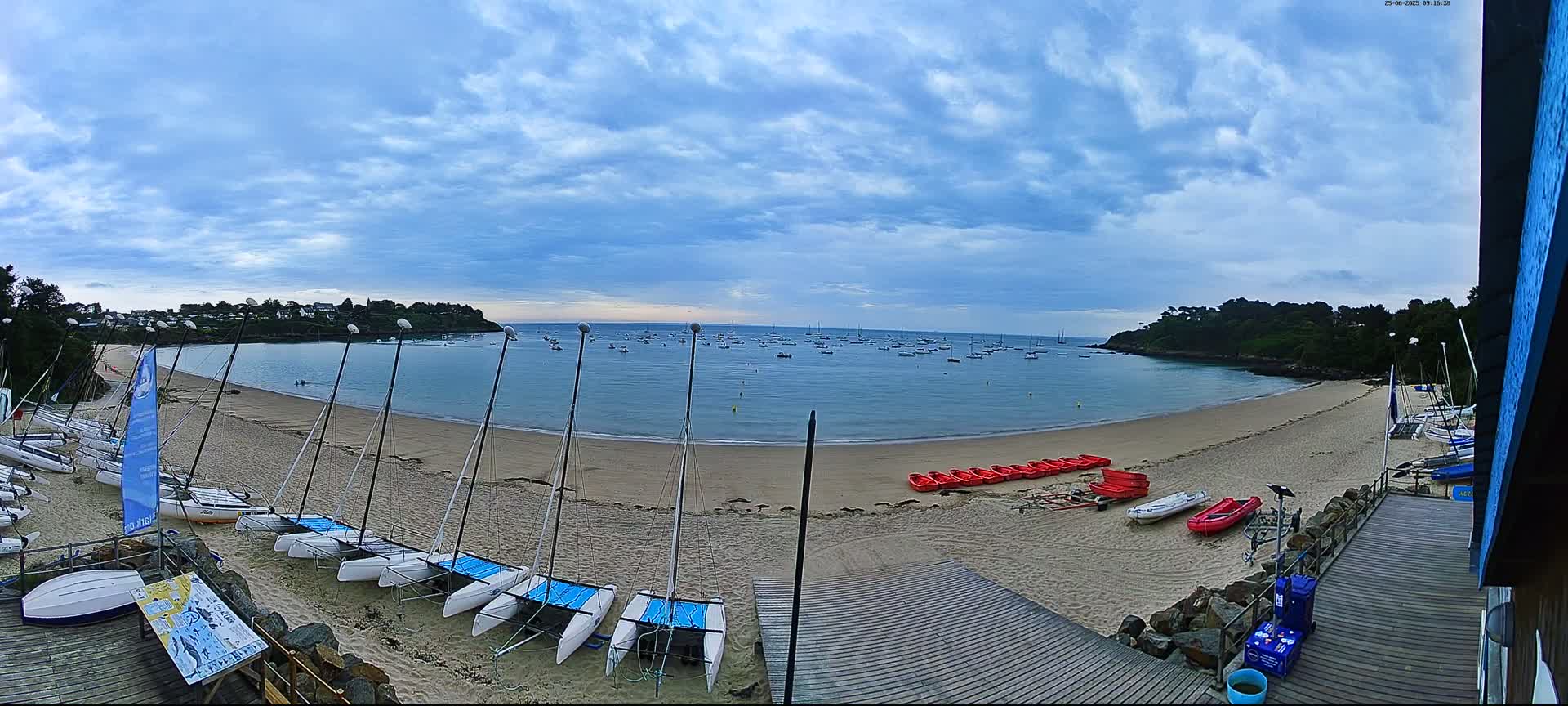 A sandy beach with numerous sailboats and small red boats on the shore, under a partly cloudy sky, overlooks a calm bay filled with more boats.