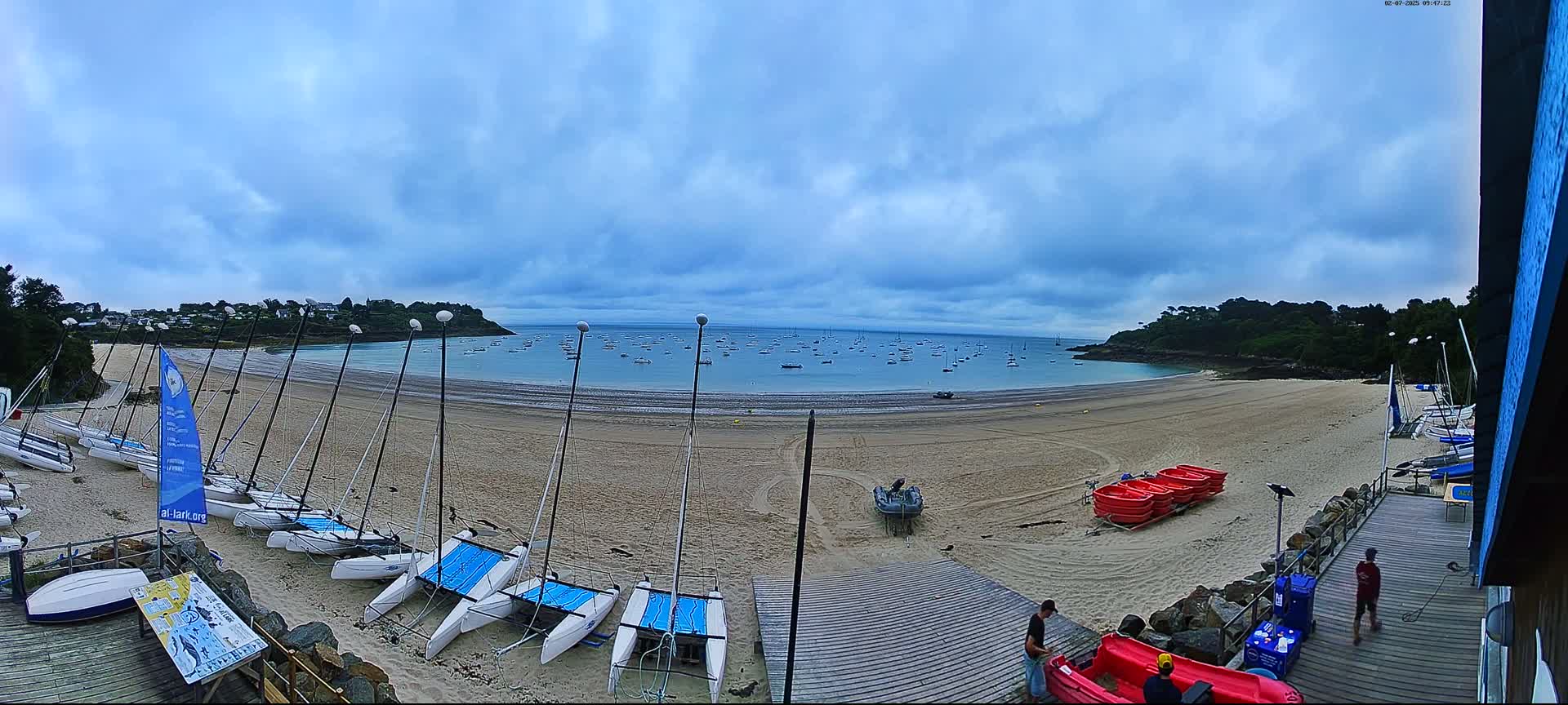 A sandy beach with numerous sailboats on the shore and many more boats in the calm water under a cloudy sky.