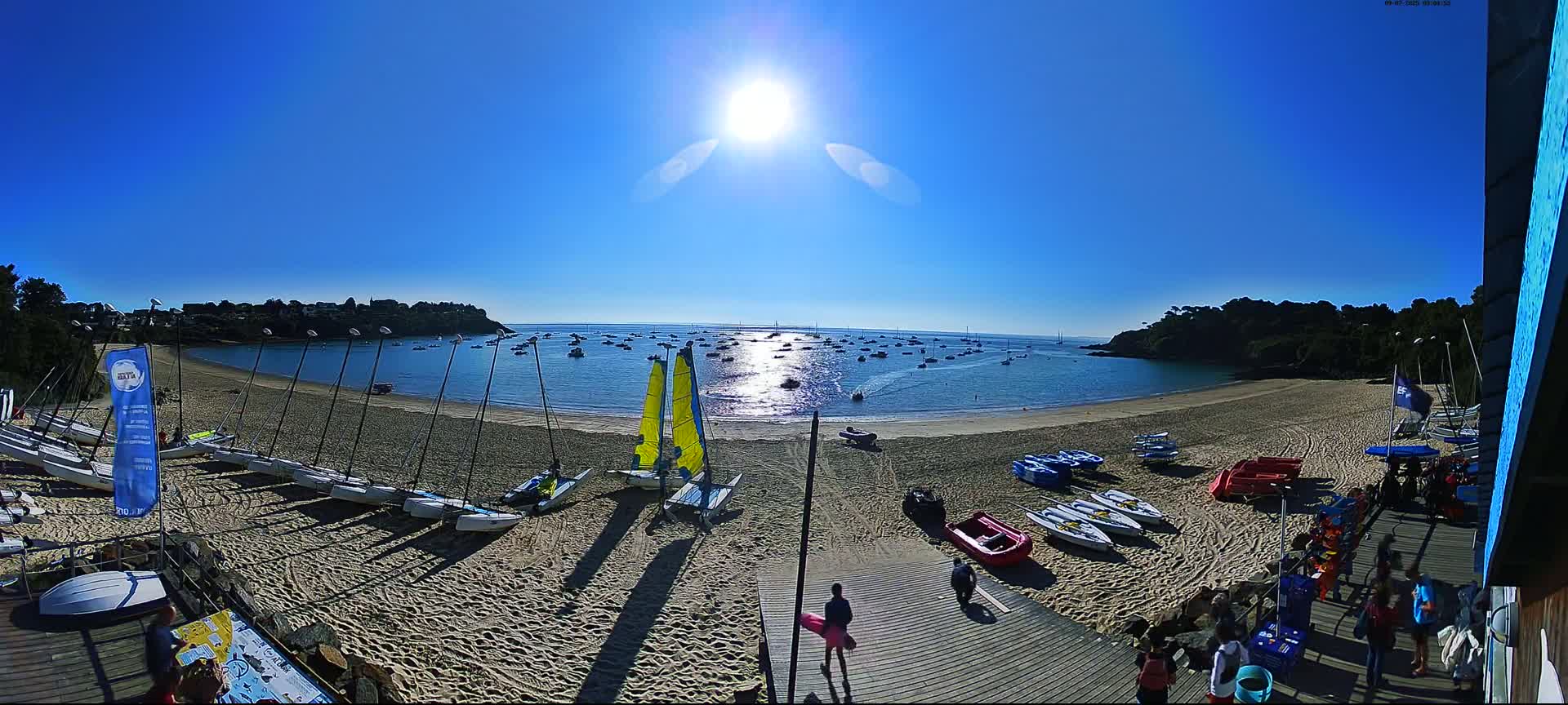 A sandy beach with numerous sailboats and small boats on the shore and in the calm, sunny water, backed by lush green hills under a bright blue sky.
