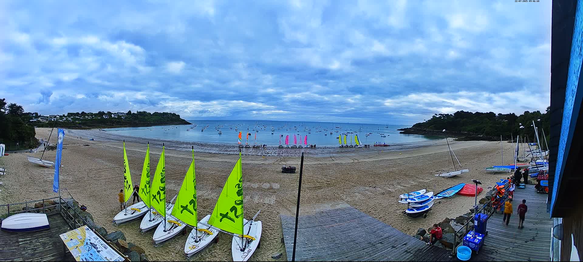 A sandy beach with numerous sailboats on the shore and in the calm water under a partly cloudy sky.