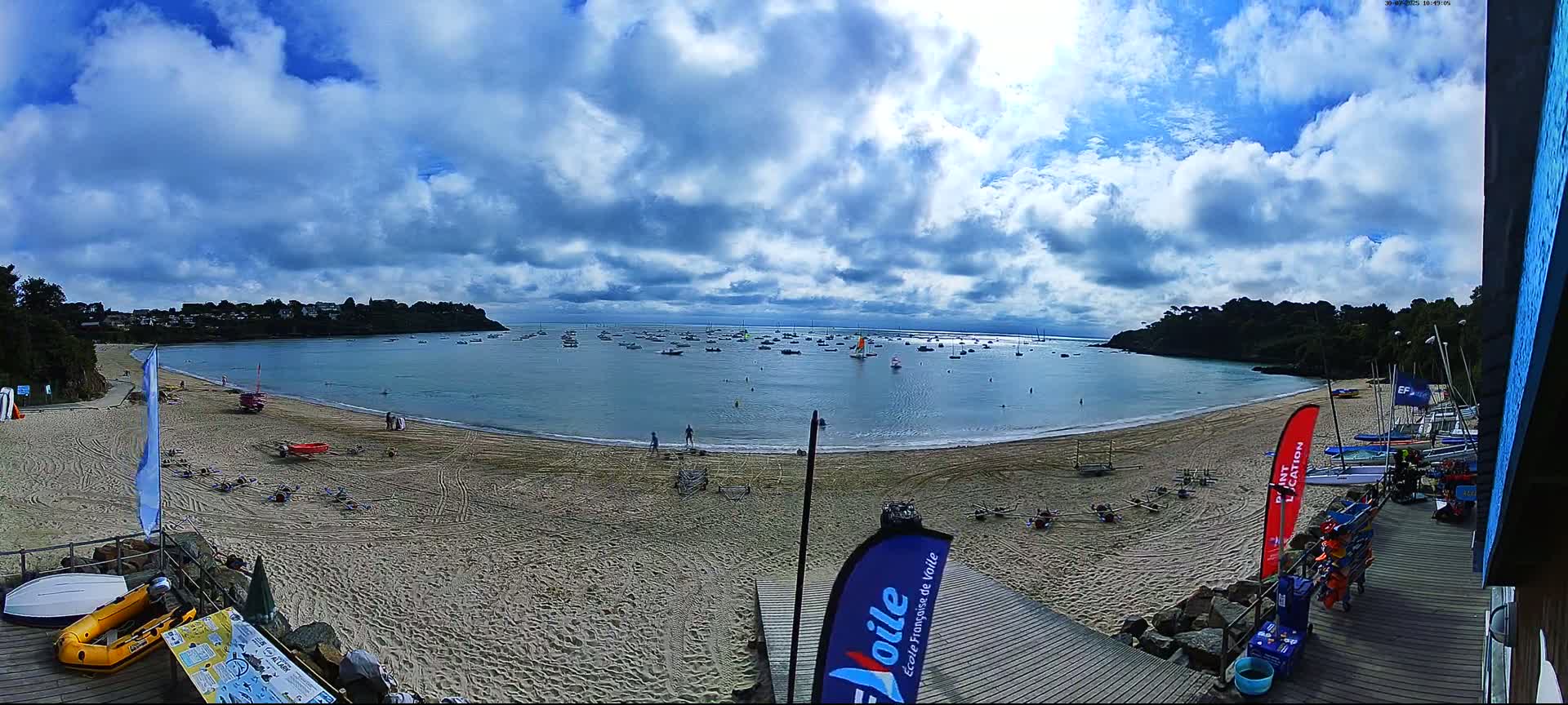 A sandy beach with numerous small boats in the calm bay under a partly cloudy sky.