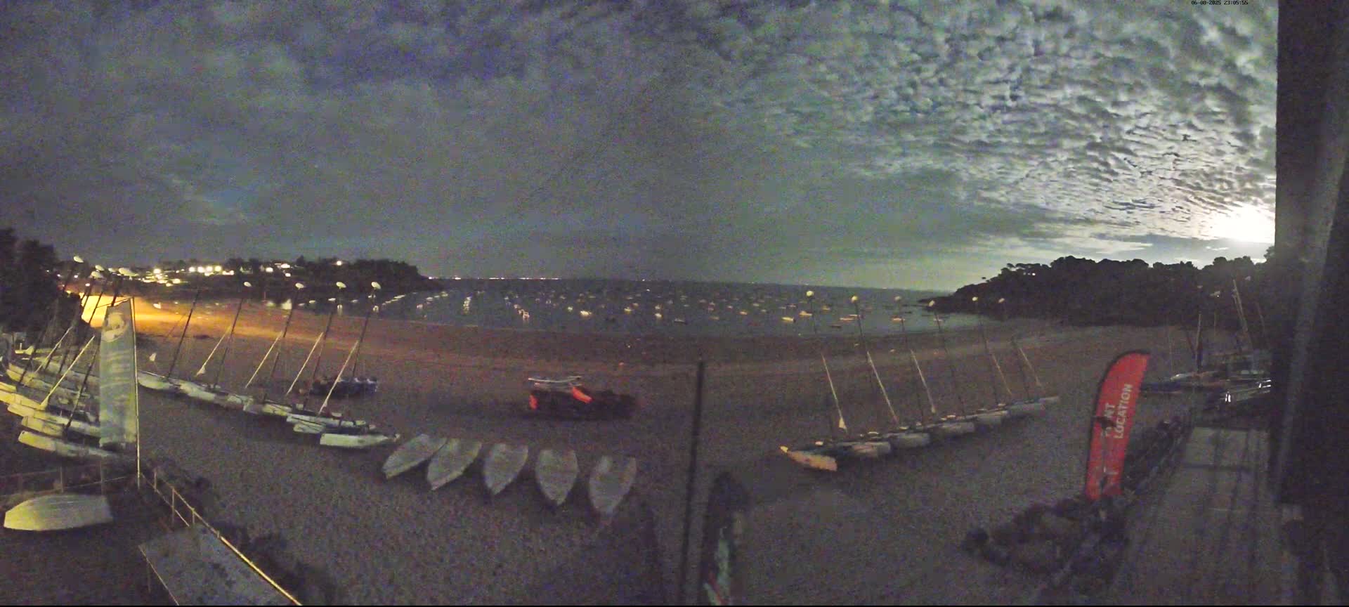 Many small sailboats are on a beach at night under a cloudy sky, with more boats visible in the water.