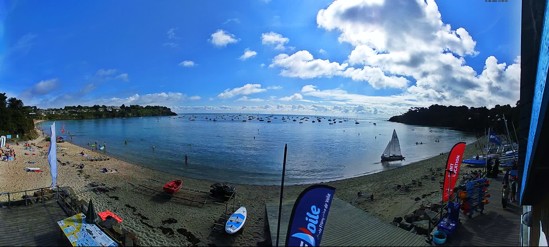 A sandy beach with sunbathers and numerous boats on a calm bay under a partly cloudy blue sky.