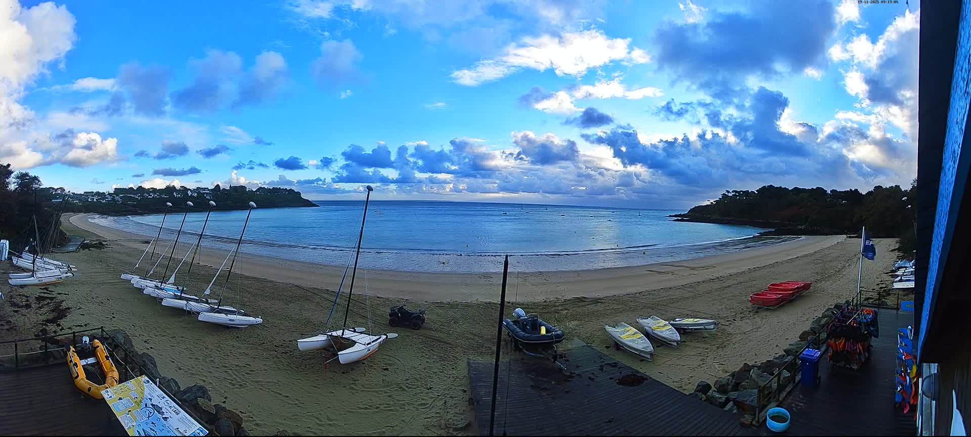 A wide, sandy crescent beach with numerous catamarans and other small boats is depicted under a partly cloudy sky featuring bright blue patches and scattered white and grey clouds, opening onto a calm blue bay flanked by forested headlands.
