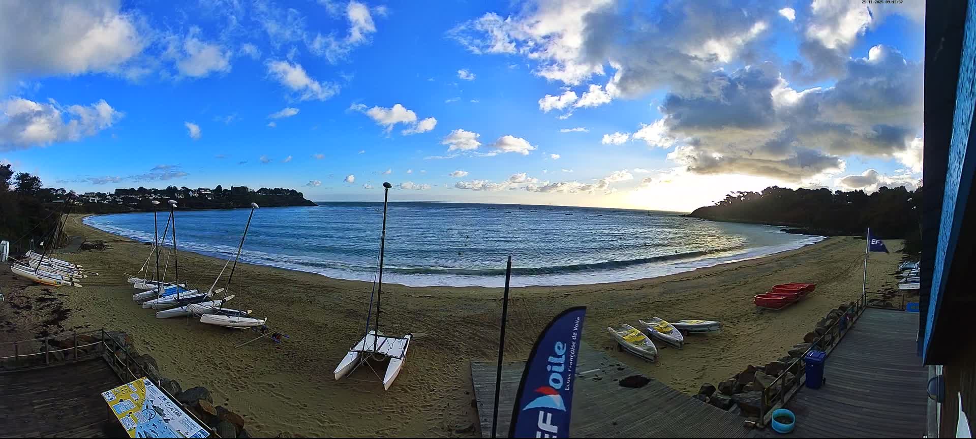 Numerous small sailboats and kayaks are scattered across a wide sandy beach that curves around a calm blue bay with gentle waves, all under a partly cloudy sky where the low sun breaks through heavier clouds on the horizon.