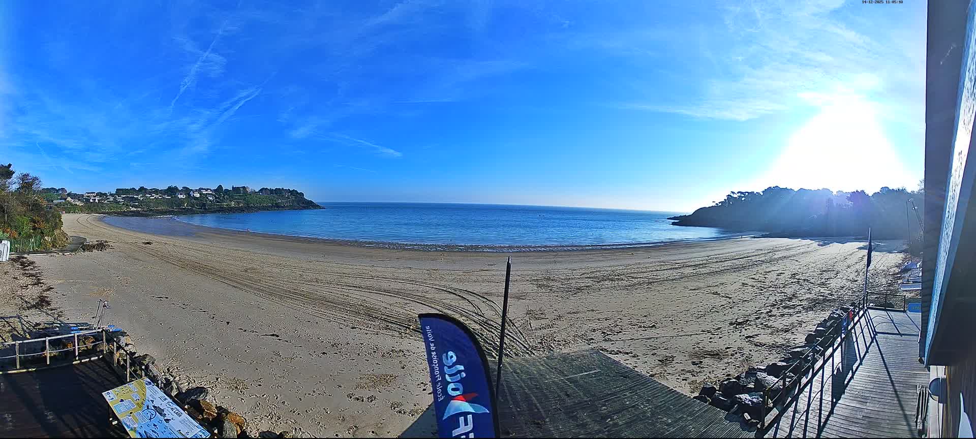 Numerous small sailboats and kayaks are scattered across a wide sandy beach that curves around a calm blue bay with gentle waves, all under a partly cloudy sky where the low sun breaks through heavier clouds on the horizon.