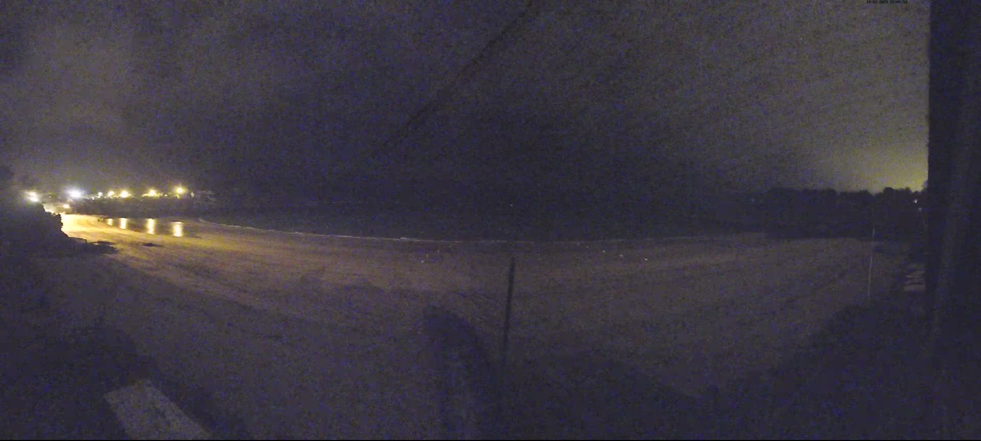 Numerous small sailboats and kayaks are scattered across a wide sandy beach that curves around a calm blue bay with gentle waves, all under a partly cloudy sky where the low sun breaks through heavier clouds on the horizon.