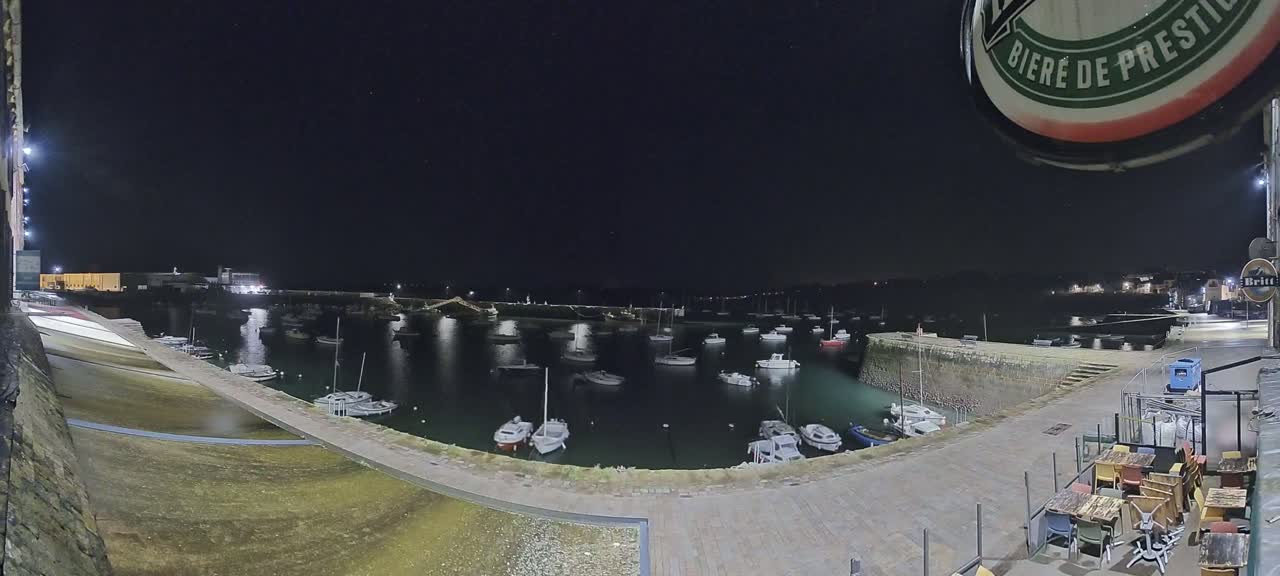 A nighttime view of a harbor filled with boats, seen from an elevated vantage point overlooking a paved area with outdoor seating.