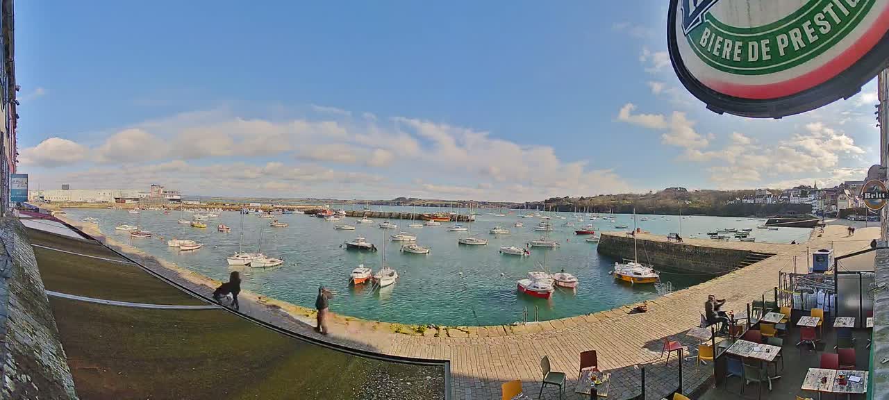 Under a mostly sunny sky with some clouds, a harbor filled with numerous sailboats and motorboats is viewed from a waterfront patio with outdoor seating.